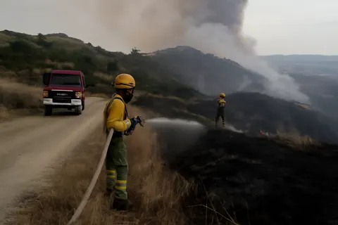 Grounded editorial scene illustrating A large wildfire in Guadalajara province, north of Madrid, has forced over 1,200 residents to evacuate as firefighters and aircraft work to contain the blaze.