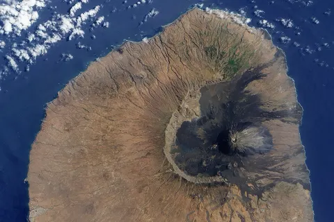 The volcano’s most distinctive feature, its nine-kilometre wide caldera, Cha Caldera, is shown in this image. The crater wall in the west towers one kilometre above the crater floor. The eastern half of the crater wall is gone, erased in a massive collapse deep in its ancient history. Evidence of the volcano’s more recent eruptive history is written on the surface of the crater as well. In the centre of the crater, a steep cone named Pico rises about 100 meters above the crater rim (more than a kilometre from the crater floor). The young peak reaches 2,829 meters above sea level, making it the island’s highest point. Dark trails of hardened lava from the volcano's most recent eruptions stream out of the crater east into the Atlantic Ocean. The volcano last erupted in 1995. The sepia stream of lava from that eruption is pooled near the crater rim west of Pico. Remarkably, the crater is inhabited. A straight road cuts between the crater wall and Pico, ending near the vent that erupted in 1995. Bright white dots on the north side of the crater are villages. Residents of the Cha Caldera evacuated during the eruption.