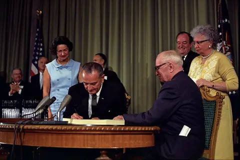 President Lyndon B. Johnson signing the Medicare Bill at the Harry S. Truman Library in Independence, Missouri . Former president Harry S. Truman is seated at the table with President Johnson. The following are in the background (from left to right): Senator Edward V. Long , an unidentified man, Lady Bird Johnson , Senator Mike Mansfield , Vice President Hubert Humphrey , and Bess Truman .