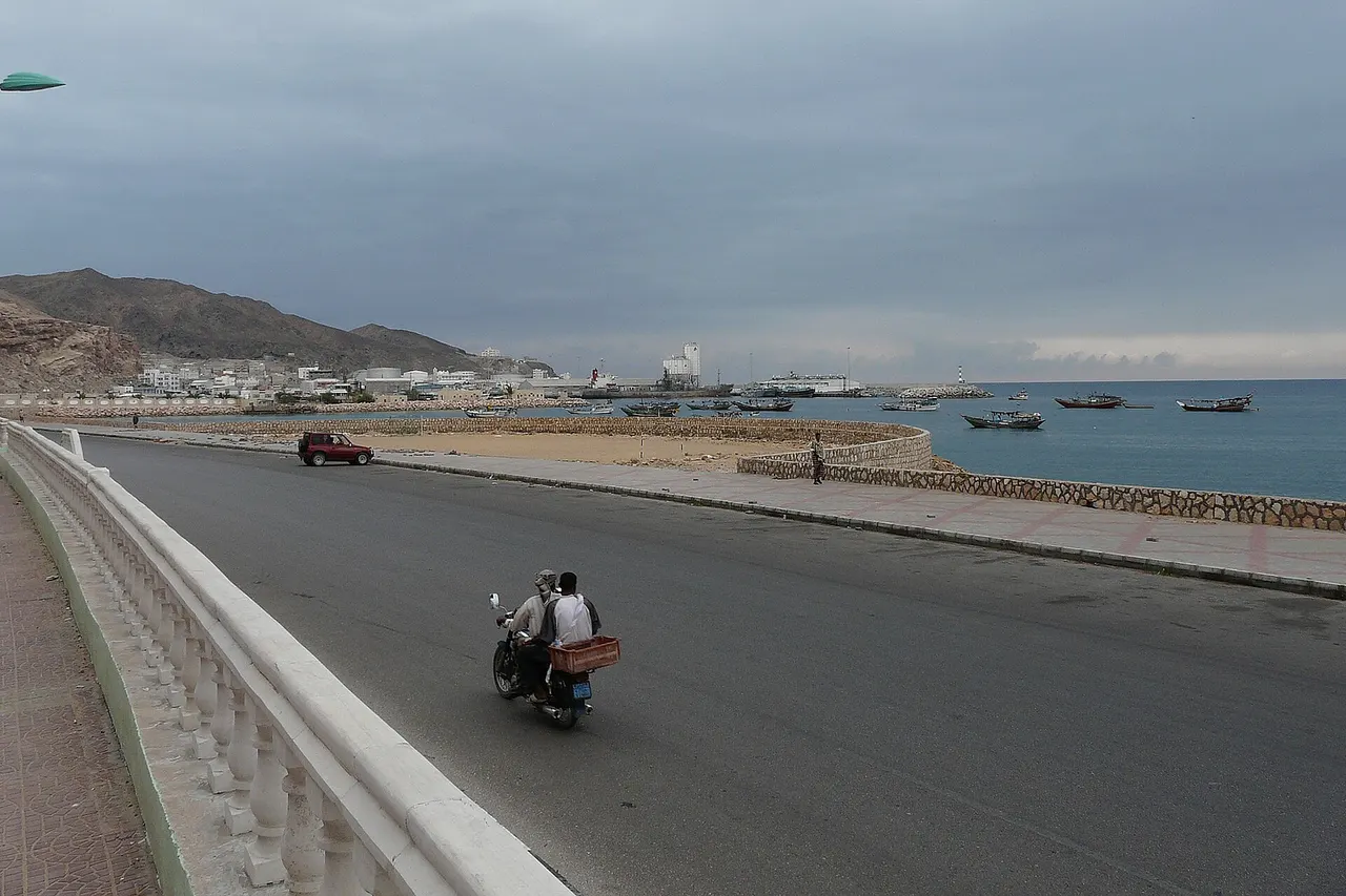 Mukalla port (Yemen). View from the route between port and town, which named Khalf Street. Cement carrier "Olympic Carrier" (gray hull, white superstructure and red funnel) is alongside. Tanks of "RAYSUT" cement company (factory in port) behind the cement carrier. Oil tanks are on the port side and behind. There are plenty of fishing vessels in port and at anchor close to the port entrance.