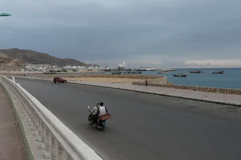 Mukalla port (Yemen). View from the route between port and town, which named Khalf Street. Cement carrier "Olympic Carrier" (gray hull, white superstructure and red funnel) is alongside. Tanks of "RAYSUT" cement company (factory in port) behind the cement carrier. Oil tanks are on the port side and behind. There are plenty of fishing vessels in port and at anchor close to the port entrance.
