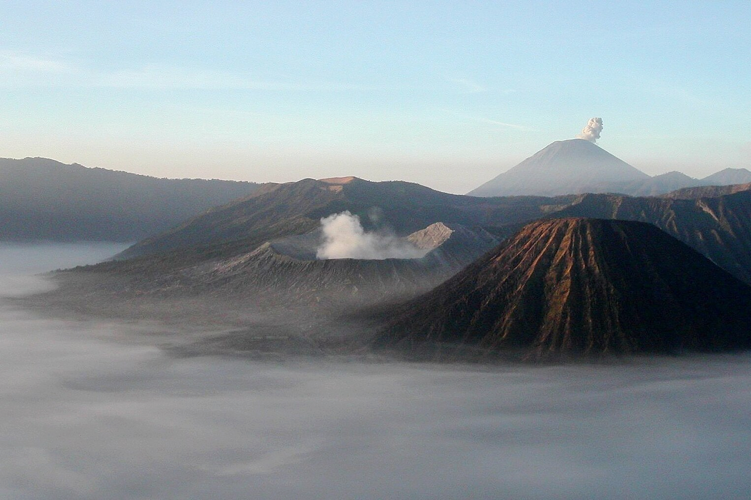 The Tengger massif in Java, Indonesia, at sunrise, showing the volcanoes Mt. Bromo (large crater, smoking), Mt. Semeru (background, smoking), Mount Batok in the foreground, and Mount Widodaren-Mount Kursi behind Mount Bromo. The early morning fog surrounds the peaks, covering a plain of finest volcanic ashes.