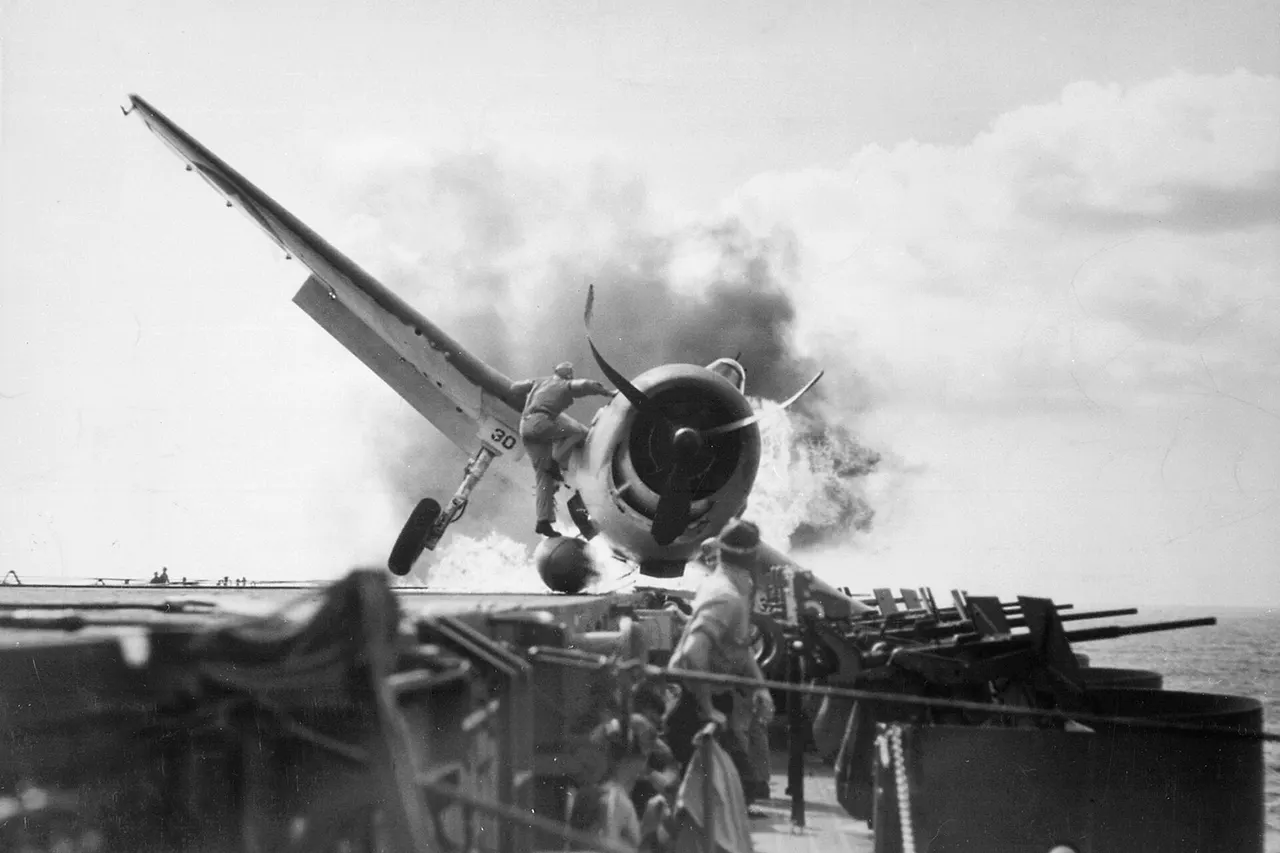 Crash landing of a U.S. Navy Grumman F6F-3 Hellcat (Number 30) of Fighting Squadron 2 (VF-2) aboard the aircraft carrier USS Enterprise (CV-6), into the carrier's port side 20mm gun gallery, 10 November 1943. Lieutenant Walter L. Chewning, Jr., USNR, the Catapult Officer, is seen climbing up the plane's side to assist the pilot from the burning aircraft. The pilot, Ensign Byron M. Johnson, escaped without significant injury. Enterprise was then en route to support the Gilberts Operation. Note the plane's ruptured belly fuel tank.