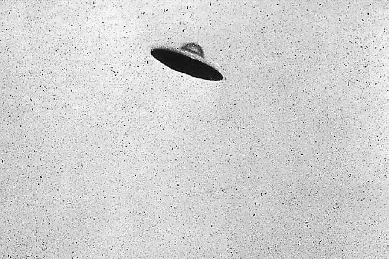 monotone non horizontal view of a supposed curvilinear centrally domed UFO relatively motionless (no blur ) above an electrocommunications pole plus cables with deciduous trees. The lighter tone of the UFO upper part relative to the lower suggests a degree of reflection of light. There is perhaps a little cloud to the right of the image, which could be part of a low-level cloud formation. A less granularitously intense band partially transverses the horizontal plane of the image, terminating leftwardly to approximately where the two phone-lines intersect. An apparent squiggle-like less intense tonal area is at the upper left of the image