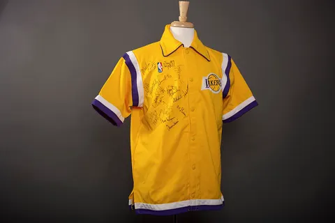 This artifact is a Los Angles Lakes Polo shirt autographed by team members: Michael Cooper, Tony Campbell, Magic Johnson, Mike Smrek, Byron Scott, Kurt Rambis, James Worthy, Kareem Abdul-Jabbar, Wes Matthews, Milt Wagner, A.C. Green, Mychal Thompson, and Jerry West. It is yellow, with purple and white stripes on short sleeves and the bottom edge of the shirt, snap button front closures. It was presented in the Oval Office. Gift of Pat Riley, Head Coach, Los Angeles Lakers.